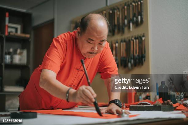 asian chinese male practising chinese caligraphy for coming chinese new year celebration home decoration purpose with prosperity and good wording by writing it on a red piece of paper
