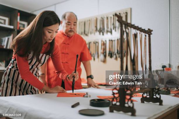 ein asiatisches chinesisches männchen übt chinesische kaligraphie für kommende chinesische neujahrsfeier zuhause dekoration zweck mit wohlstand und gute formulierung, indem sie es auf einem roten stück papier und lehre seine daugther - 55 year old male stock-fotos und bilder