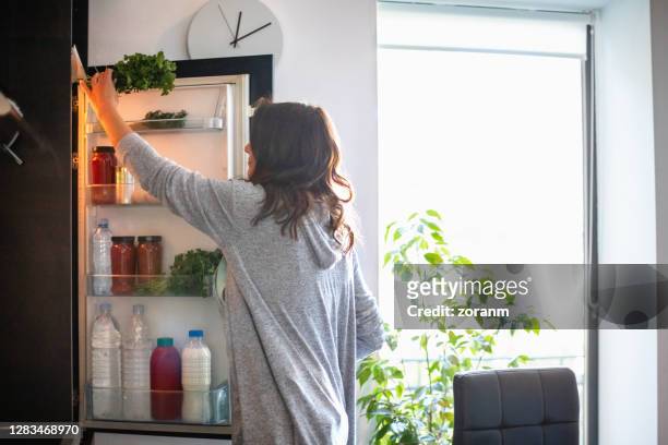 woman by the open fridge taking a bunch of parsley - refrigerator stock pictures, royalty-free photos & images