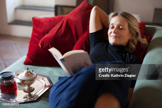 beautiful mature woman relaxing on the sofa with cup of tea and a book - reading stock-fotos und bilder