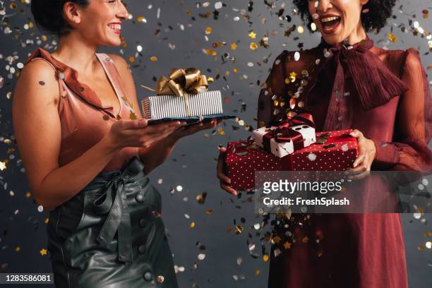 party time: twee elegante vrouwen uitwisselen van kerstcadeaus op een feestje, confetti flying all around them - christmas party stockfoto's en -beelden