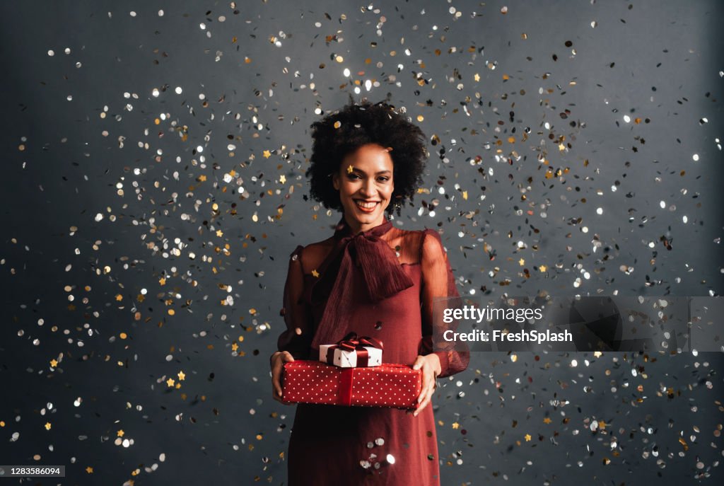 Frohe Weihnachten: eine glückliche afroamerikanische Frau in einem roten festlichen Kleid hält Weihnachtsgeschenke mit Konfetti fliegen rundum, ein Porträt