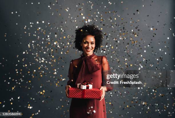 merry christmas: a happy african american woman in a red festive dress holding christmas presents with confetti flying all around, a portrait - african ethnicity christmas stock pictures, royalty-free photos & images