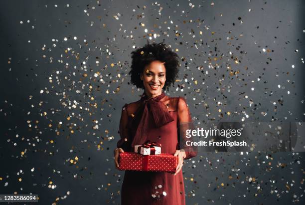 merry christmas: a happy african american woman in a red festive dress holding christmas presents with confetti flying all around, a portrait - red dress imagens e fotografias de stock