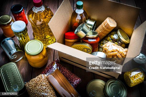 caja de cartón llena de alimentos no perecederos sobre mesa de madera. vista de ángulo alto. - emergencies and disasters fotografías e imágenes de stock