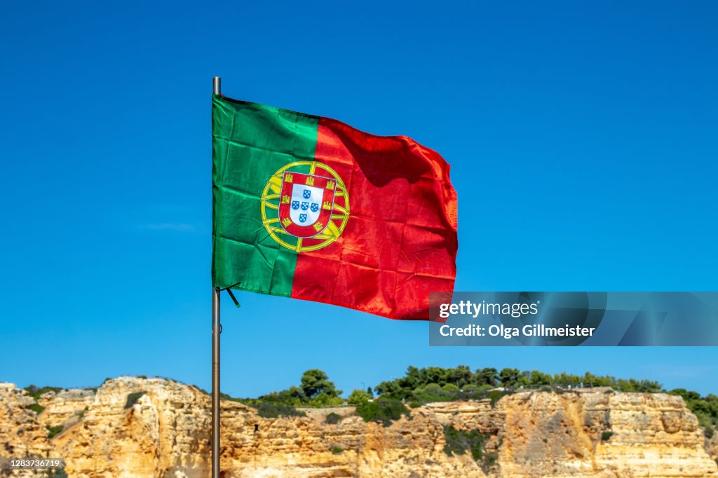 Selective focus on waving flag of portugal in front of typical yellow rock formation of algarve and blue sky. National portugal flag with space.