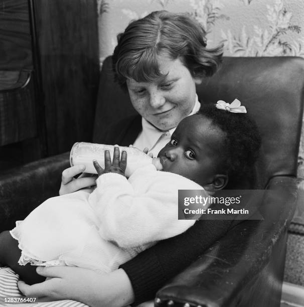 The mixed race Long family of Olyffe Avenue in Welling, Kent, July 1960. Twelve-year-old Joan helps feed her adopted sister, baby Kim.