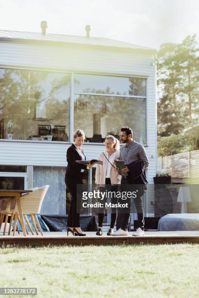 real estate agent showing magazine of property to couple against new house - estate agent stock pictures, royalty-free photos & images