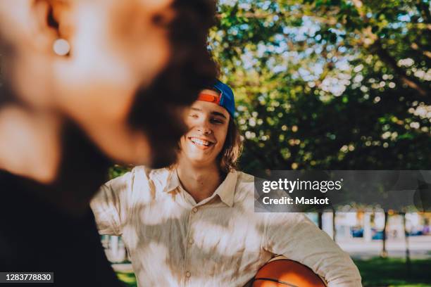 portrait of smiling male with friend holding soccer ball in park - teenage boy in cap posing stock pictures, royalty-free photos & images