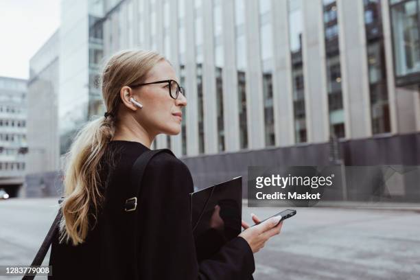 rear view of businesswoman with in-ear headphones holding file in city - ohrhörer stock-fotos und bilder