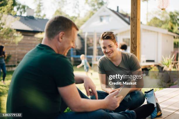 side view of smiling male showing digital tablet to homosexual partner while mothers and son playing in backyard - son in law stock-fotos und bilder