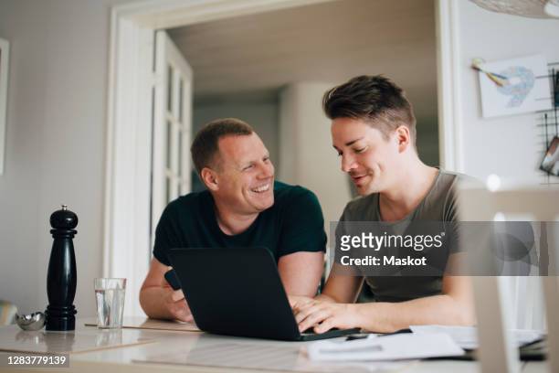 male talking to smiling partner while using laptop over dining table at home - mid adult couple stock pictures, royalty-free photos & images