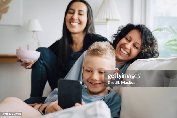 smiling lesbian couple looking at son using mobile phone on sofa at home - lesbian mom stock pictures, royalty-free photos & images