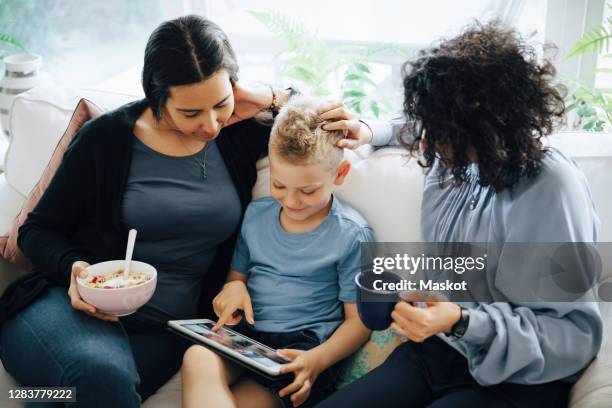 son using digital tablet on sofa by lesbian mothers at home - lesbica foto e immagini stock