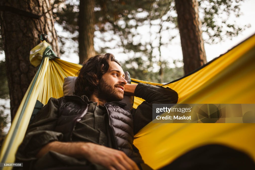 Smiling man looking away while lying over hammock in forest