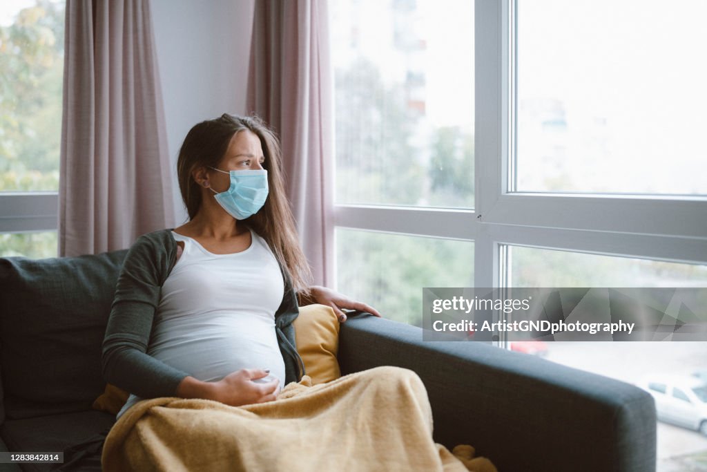 Mujer embarazada preocupada con mascarilla protectora en casa.