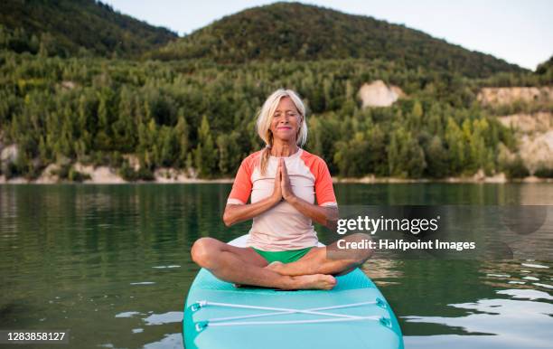 active senior woman on paddleboard on lake in summer, doing yoga. - practioners enjoy serenity of paddleboard yoga stockfoto's en -beelden