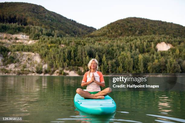 active senior woman on paddleboard on lake in summer, doing yoga. - paddle board stock pictures, royalty-free photos & images