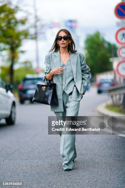 Guest wears sunglasses, earrings, a green oversized blazer jacket, three-piece suit, flared pants, a black leather Hermes bag, outside Hermes, during...