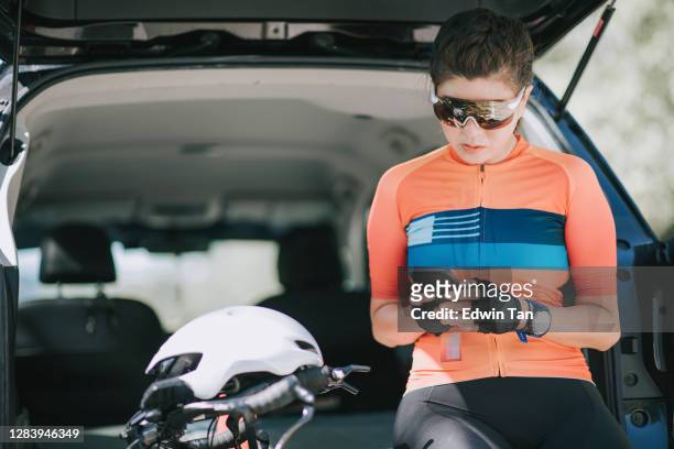 asian chinese female cyclist sitting at the suv car trunk using phone replying message taking a break - camisola de ciclismo imagens e fotografias de stock