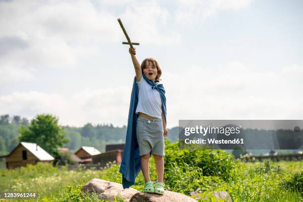 playful boy wearing cape holding toy sword while standing on rock against sky - swords stock-fotos und bilder