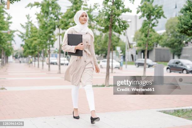 muslim teenage girl holding folder while walking on sidewalk in city - islamic action front stock pictures, royalty-free photos & images