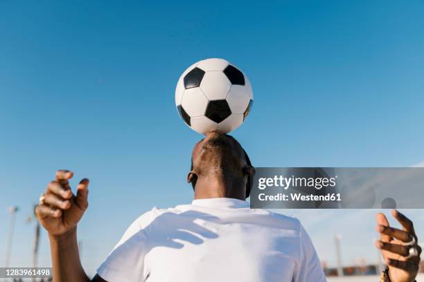 close-up of young man balancing soccer ball on head against blue sky - football ball stock-fotos und bilder