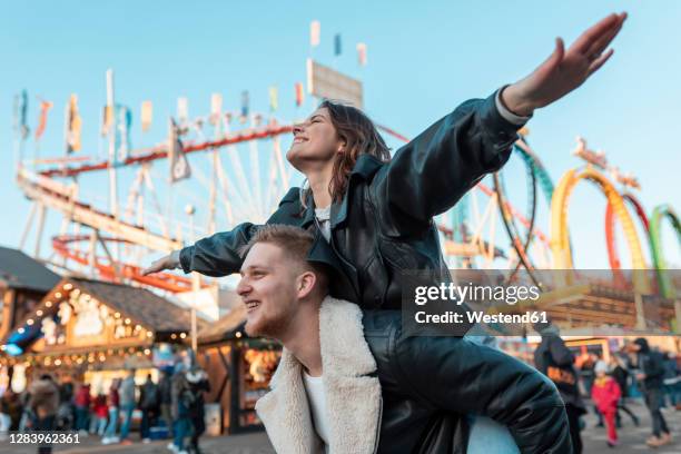 happy man piggybacking carefree girlfriend while standing at amusement park - pretpark stockfoto's en -beelden