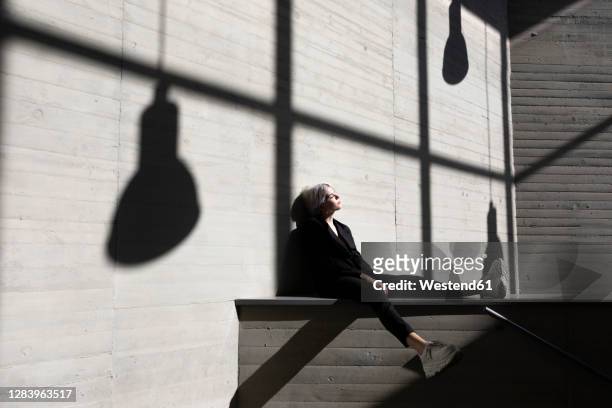 businesswoman wearing elegant suit sitting on retaining wall with sunlight and shadow in background - shadow wall stock-fotos und bilder
