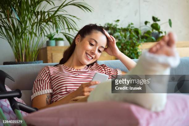 mujer joven con la pierna rota usando el teléfono inteligente - pierna fracturada fotografías e imágenes de stock