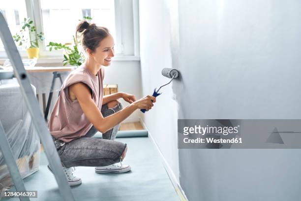 young woman painting wall with paint roller while crouching at home - painting stock pictures, royalty-free photos & images