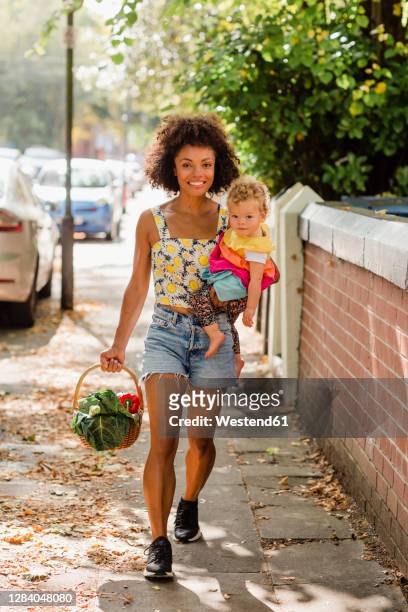 smiling mother carrying baby and vegetable basket while walking in city - baby on the move stock pictures, royalty-free photos & images