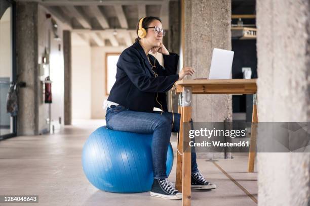 businesswoman wearing headphones using laptop while sitting on fitness ball at office - fitness ball imagens e fotografias de stock