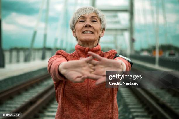 reife frau streckt arme auf einer brücke immer bereit für das laufen - old woman running stock-fotos und bilder