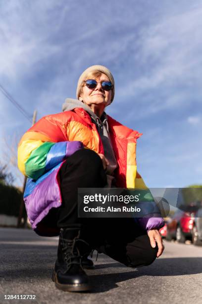 modern grandmother in rainbow coat posing in the middle of the street. - gay flag stock-fotos und bilder