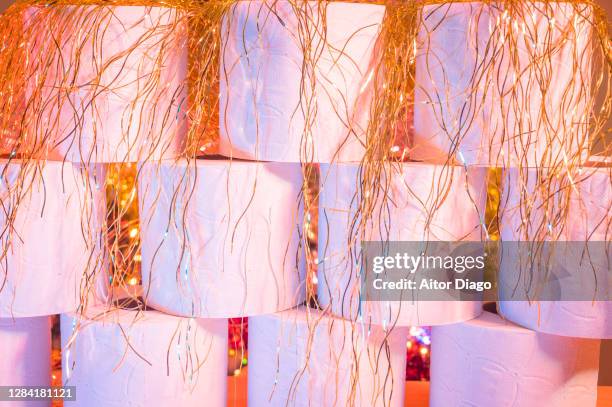 close-up of  toilet papers roles with goldfish thin garlands. in the background and out of focus, there is a christmas tree decorated with colorful lights. - toilet paper tree 個照片及圖片檔