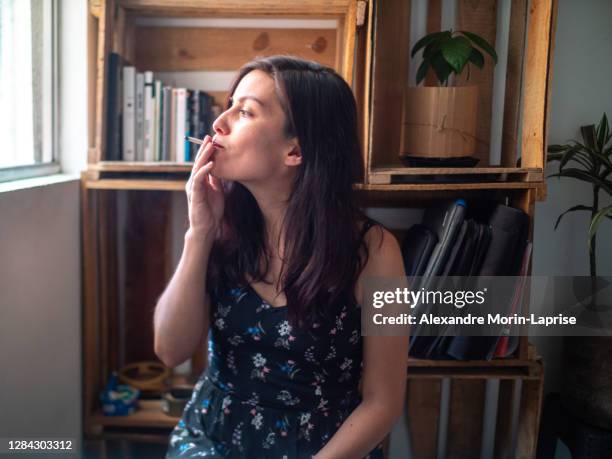 young attractive hispanic woman in blue dress smokes hand rolled cigarrette / joint by apartment window - femme et fumeuse photos et images de collection