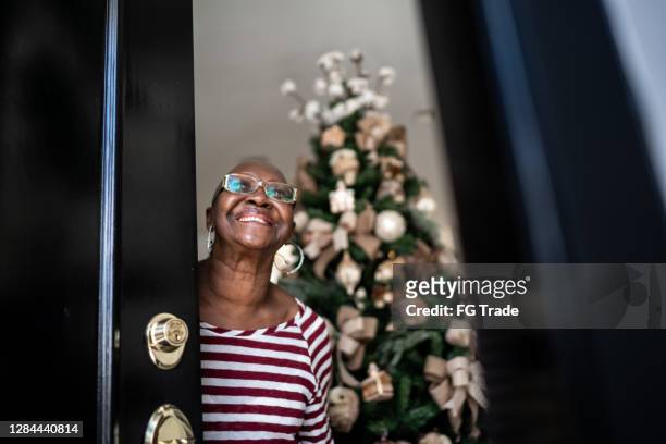 gelukkige hogere vrouw die huisdeur, kerstboom op de achtergrond opent - homeowners decorate their houses for christmas stockfoto's en -beelden
