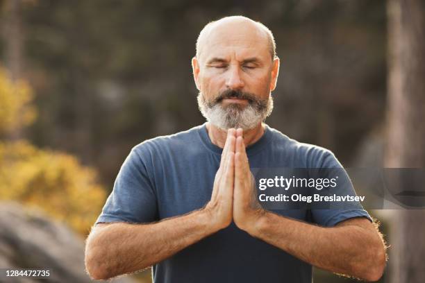 close up portrait of old gray-haired senior man with beard practicing yoga outdoors, meditating and praying - guru stock-fotos und bilder