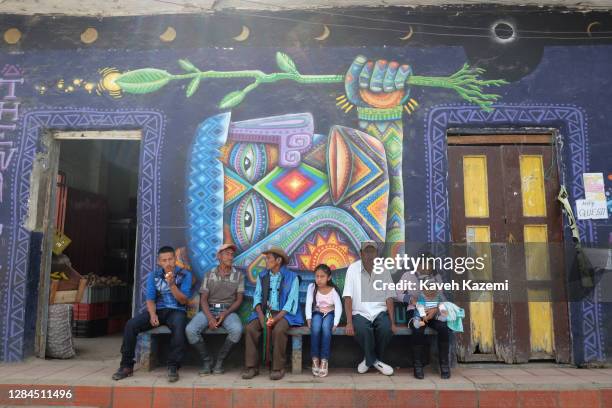 Indigenous Páez people, also known as the Nasa sit in front of a mural with iconography of their culture outside the local store on the side of the...