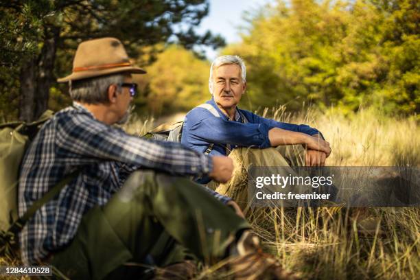 two senior men sitting on grass in forest and talking while taking a break during hiking - flannel stock pictures, royalty-free photos & images