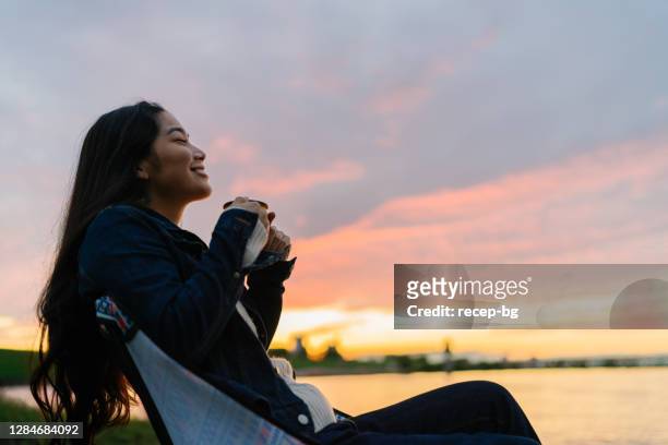joven mujer disfrutando de bebida caliente en la naturaleza durante la puesta del sol junto al lago - japon fotografías e imágenes de stock