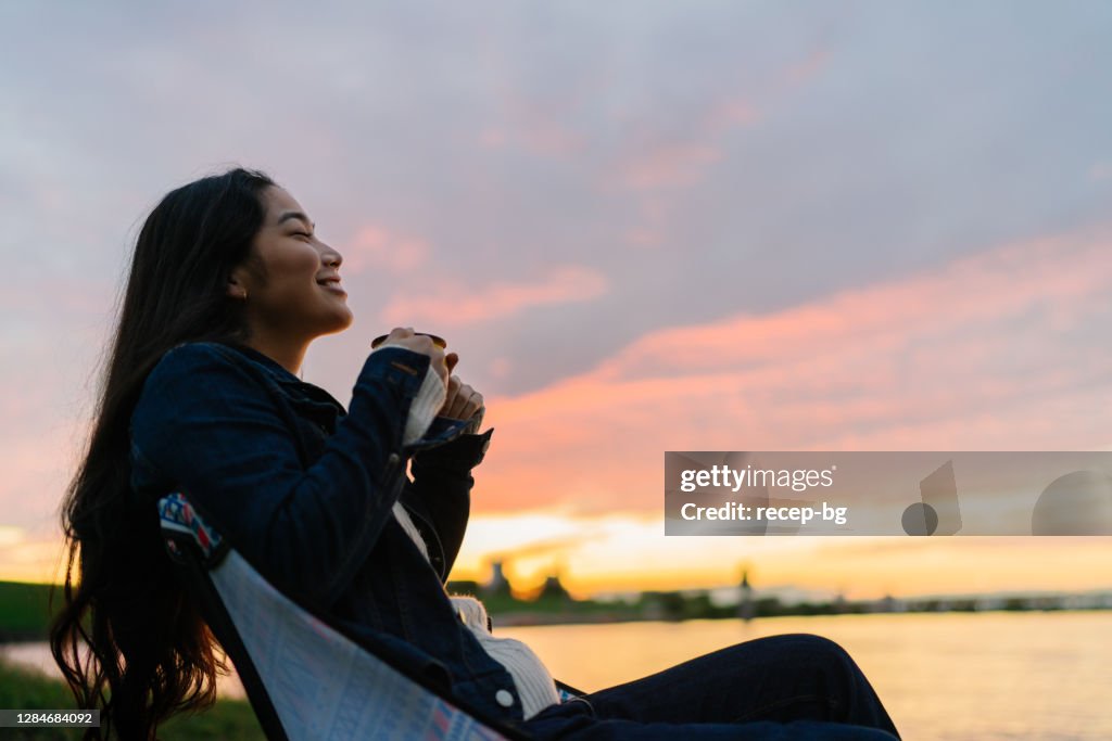 Young woman enjoying hot drink in nature during sunset by lake