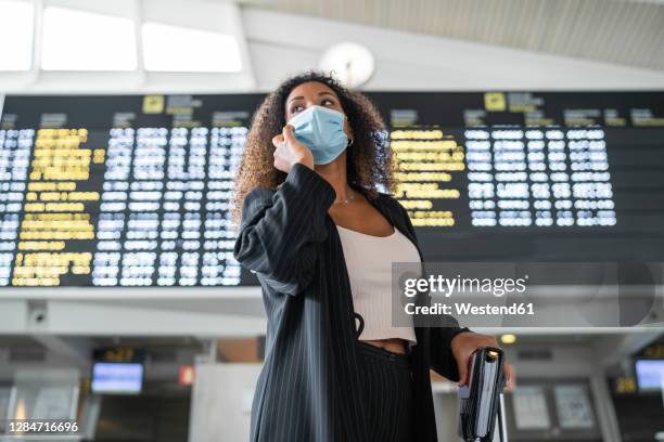 woman wearing protective face mask talking on smart phone at airport - departure board front on fotografías e imágenes de stock