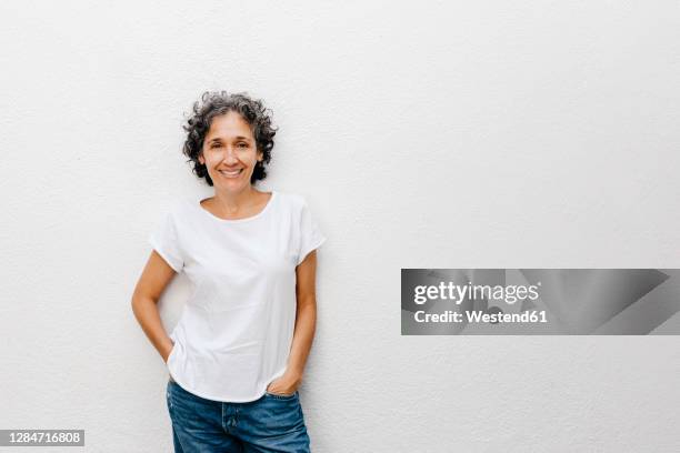 smiling mature woman with short hair standing against white wall - european woman stock-fotos und bilder