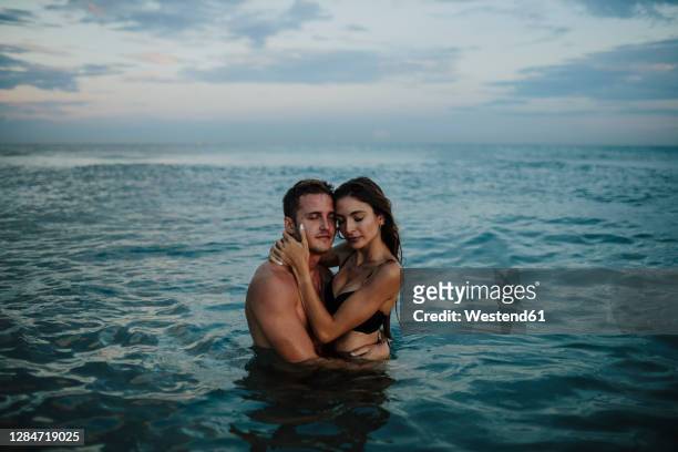 couple with eyes closed standing in water at beach during sunset - bathing in sunset stock pictures, royalty-free photos & images