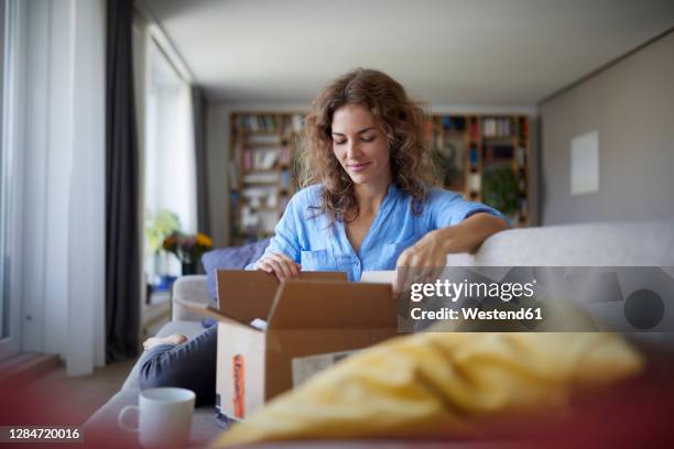 woman opening box while sitting on sofa at home - boxseat stock-fotos und bilder