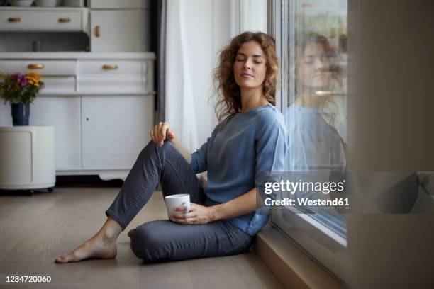 mid adult woman with coffee cup sitting while leaning on glass door at home - relief emotion stock pictures, royalty-free photos & images