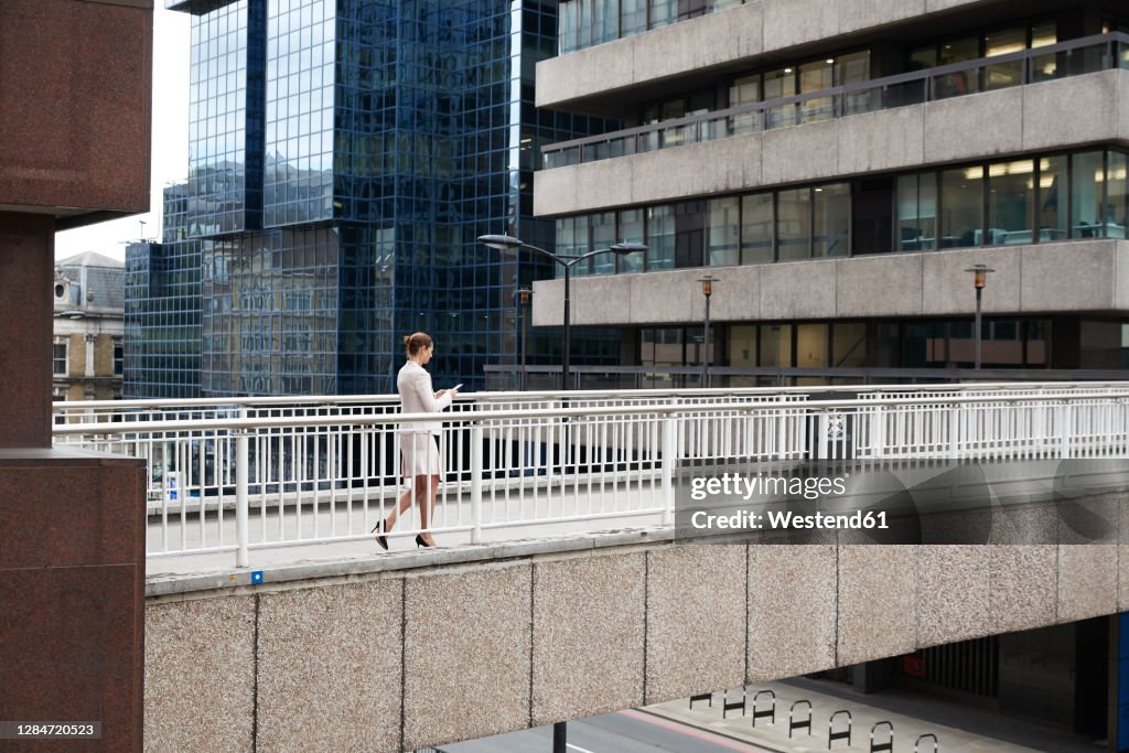 Businesswoman using smart phone while walking on footbridge at downtown district