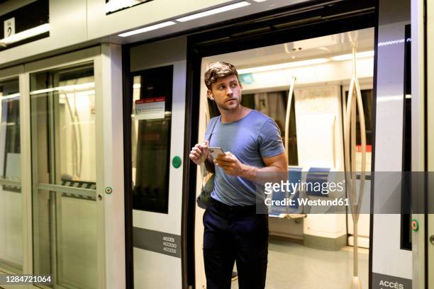 handsome young male commuter looking away while disembarking from subway train at station - disembarking stock-fotos und bilder